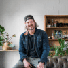 man wearing a denim jacket and baseball hat sitting on a couch