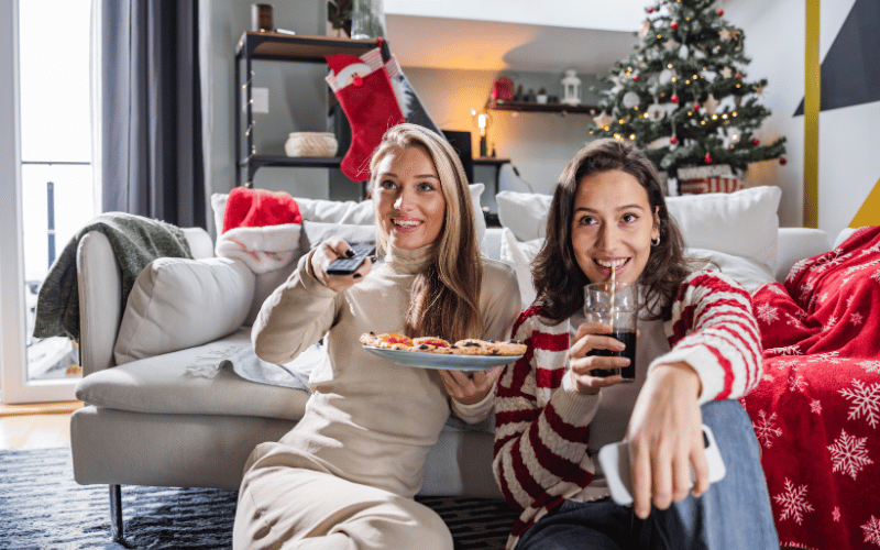 Two 30-something women eating Christmas cookies and watching holiday movies in front of the couch