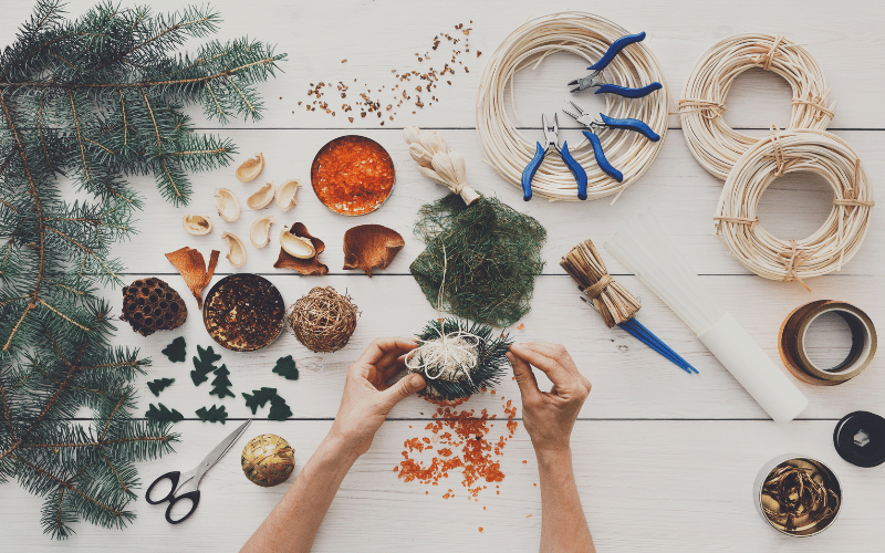 Hands painting and tying together DIY Christmas Decorations with twine, evergreen branches and pine cones