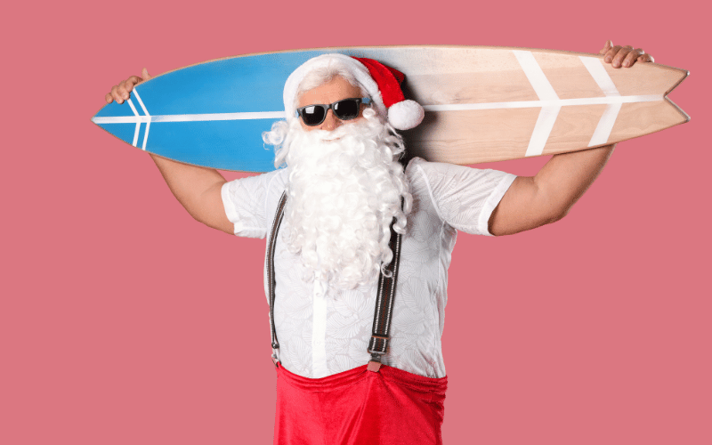 30-something man dressed as beach santa weawring sunglasses, with red pants and suspenders over white button up holding blue adn brown surf board in front of pink background