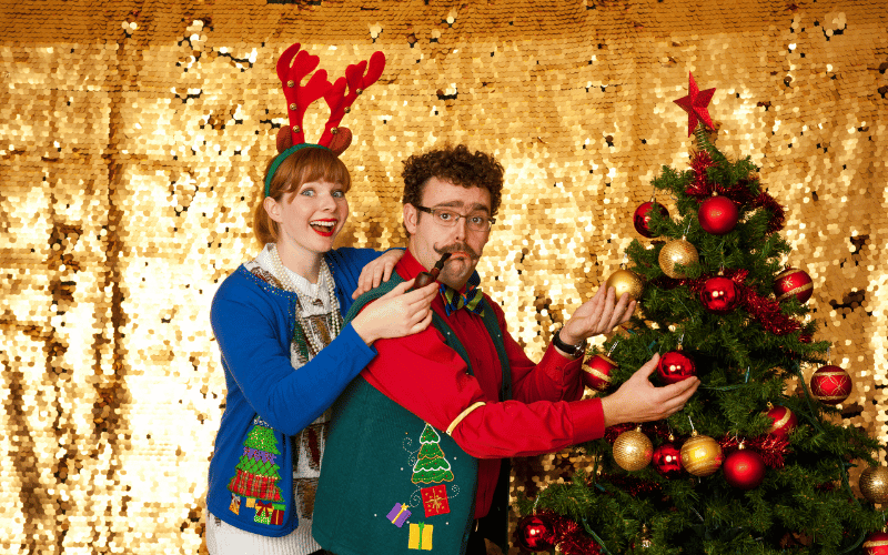 30-something couple with the girl wearing a dark blue ugly Christmas cardigan and reindeer antlers, and her boyfriend wearing  a green ugly sweater vest posing with a Christmas Tree over a gold backdrop