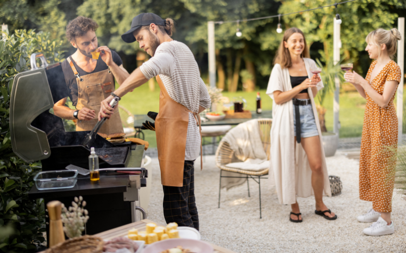 Group of young friends enjoying a backyard BBQ with one man BBQing and the others enjoying an adult beverage
