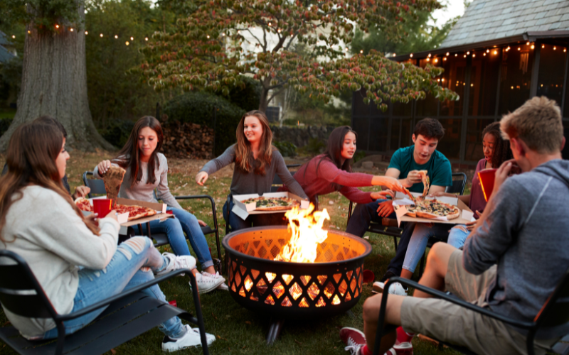 Group of 20-30-somethings gathered around a fire outdoors sharing pizza and smiling 