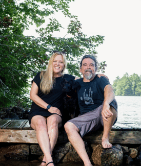 Ashley Morrill and Ryan Eldridge sitting together smiling on a dock on a lake in front of trees