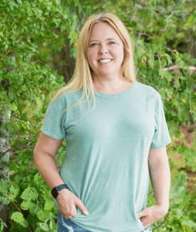 Ashley Morrill from Maine Cabin Masters smiling for camera in front of greenery wearing pale green tshirt