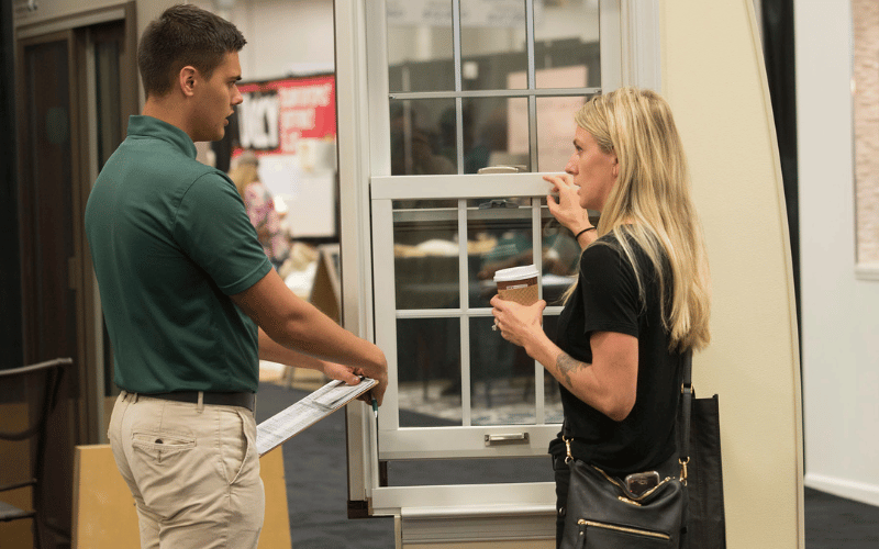 Young male employee wearing green shirt taking to blonde woman with coffee about her windows at the home show