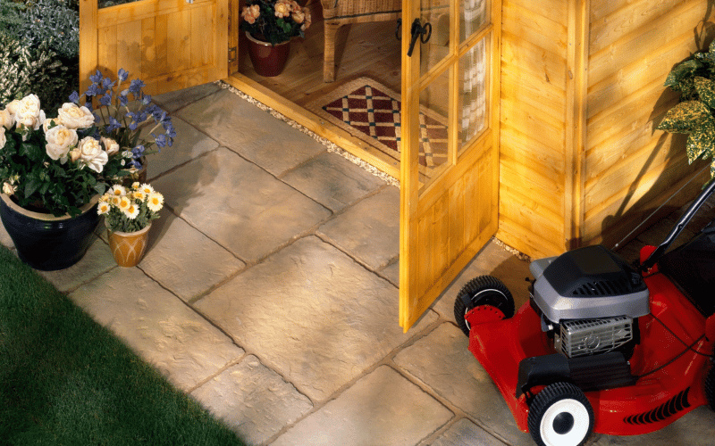 birds eye view of red lawnmower next to wooden outdoor shed with doors open with clean patio stones, spring white and purple flowers on a sunny day