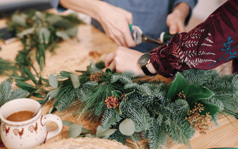 Two people trimming pine decorations in order to make a DIY wreath on wooden table with coffee