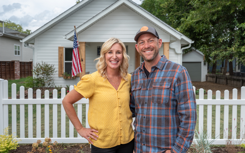 Jenny Marrs wearing yellow button up shirt with her husband, Dave Marrs, in front of house and white picket fence