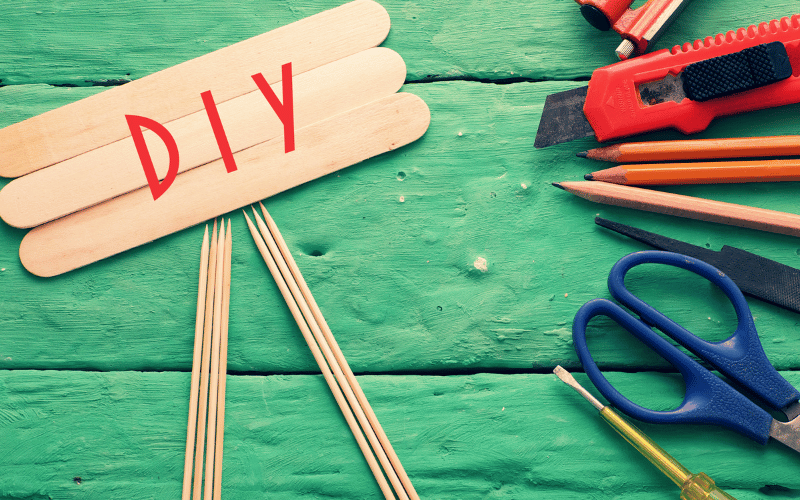 DIY written on popsicle sticks in red beside scissors, pensils and exacto blade in front of green painted wood background