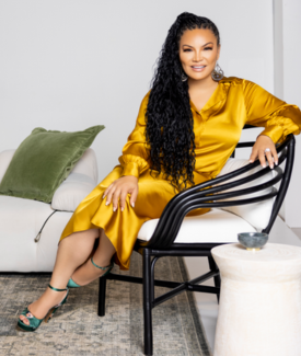 Egypt Sherrod wearing a shiny yellow dress and green shoes sitting on a black designer chair smiling