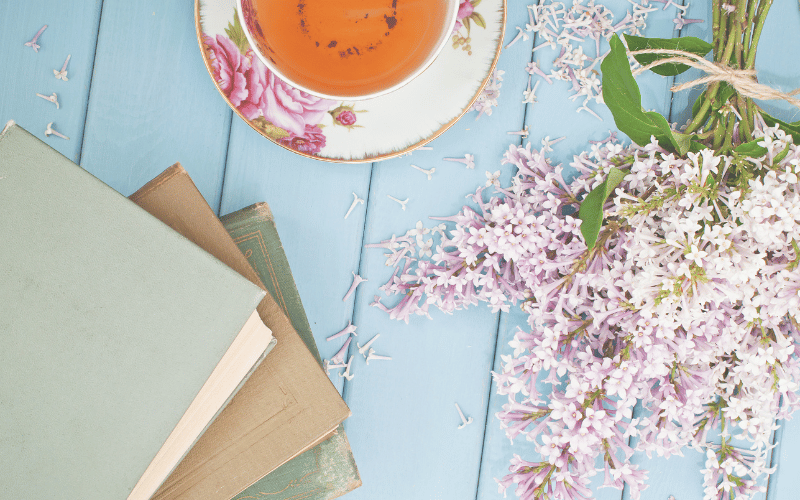 Small stack of pastel covered books next to tea and pink flowers over light blue painted deck