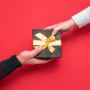 Bright red background with female hand wearing white sweater giving black and gold wrapped gift to male hand below.