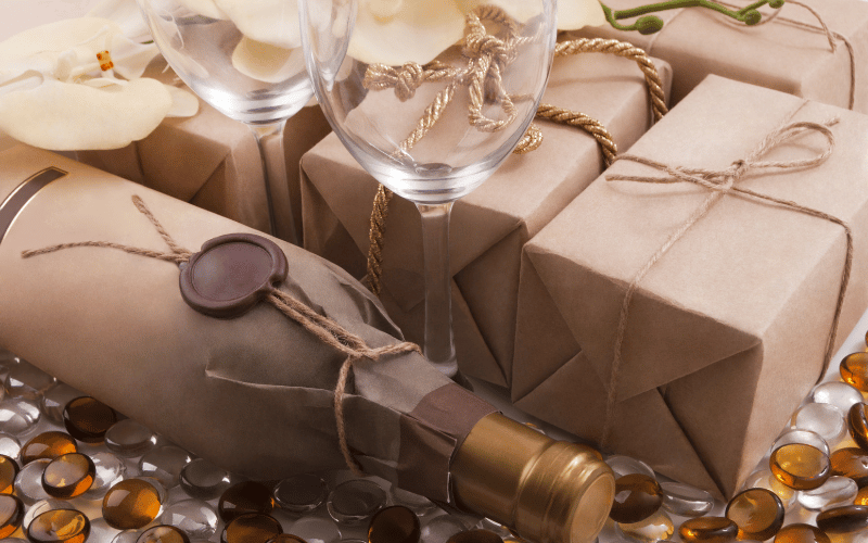 Brown paper wrapped wine bottle beside presents wiith rope bow and wine glass on table
