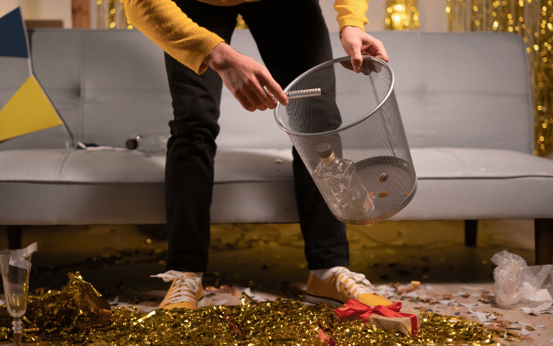 close up of person wearing yellow shirt and shoes and black pants picking up garbage off the floor and putting it into basket after party