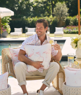 Craig Conover wearing khakis and a pink Hawaiian shirt clutching white and pink flamingo pillow and holding a drink sitting in a backyard patio chair by the pool