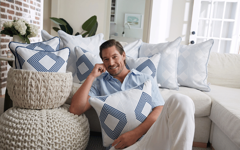 Craig Conover leaning against white couch holding blue and white pillows smiling while touching his right cheek with his right hand