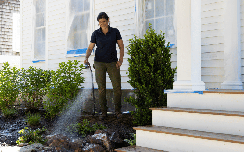 woman standing on the porch of a large white color house with garden plants