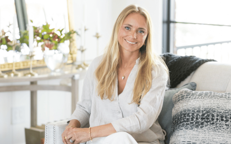 a blonde woman wearing a white silk shirt sitting on a white couch in a living room 