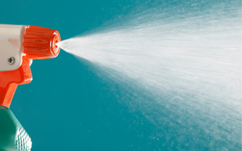 green spray bottle with orange nozzle and handle spraying bleach over blue background