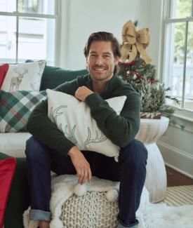 Craig Conover wearing green sweater hugging white reindeer pillow sitting on couch in front of Christmas Tree