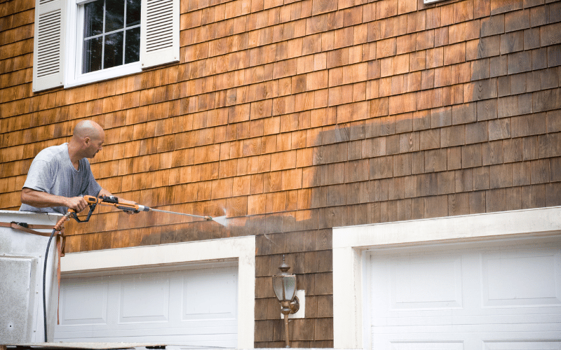 Bald white man wearing grey tshirt power washing a brown brick house