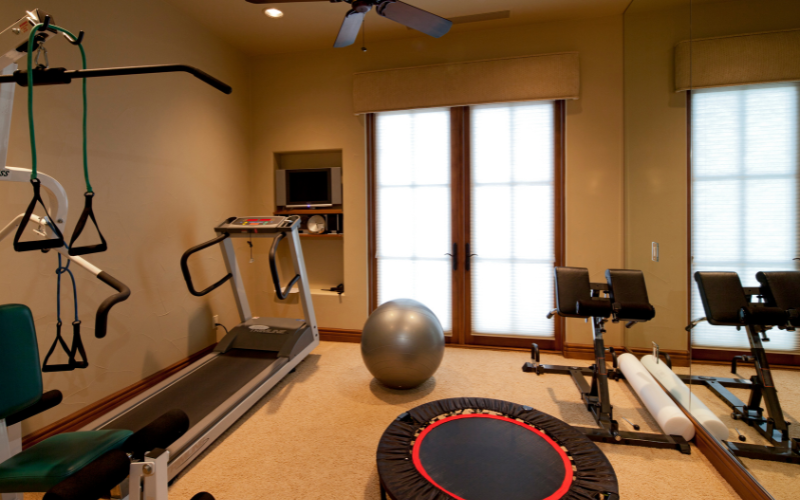 Home gym with treadmill, trampoline, exercise bands, grey pilates ball, and squat machine in front of double glass patio doors 