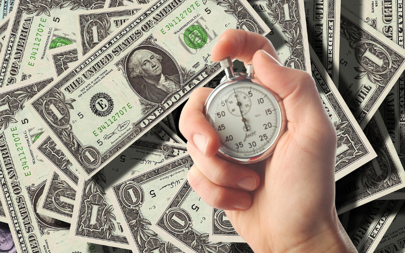 pile of American dollar bills with small white hand holding small silver timer