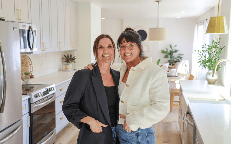 Leslie Davis wearing black blazer next to sister, Lyndsay Lamb wearing white sweater, embracing and smiling in modern kitchen