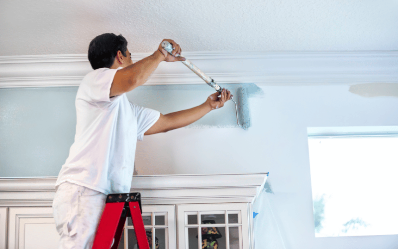 man wearing white stands on red ladder and uses roller to paint walls pale blue-grey