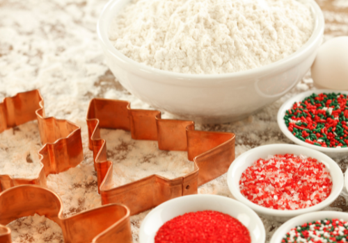 White bowl of flour red metal cookie cutters and bowls of sprinklers on flour covered countertop