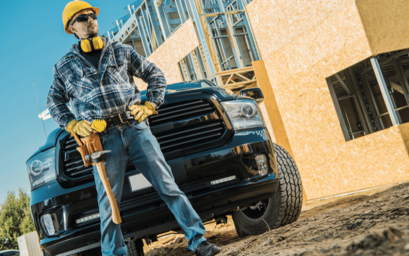 Middle age white male contractor wearing yellow hardhat and yellow noise cancelling headphones around neck over blue paid shirt with sledge hammer on belt standing next to under contraction home in front of blue truck