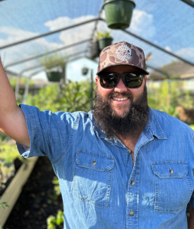 Destin-Noak-Texas-Garden-Guy-Smiling-In-Greenhouse wearing blue denim button up and camo hat