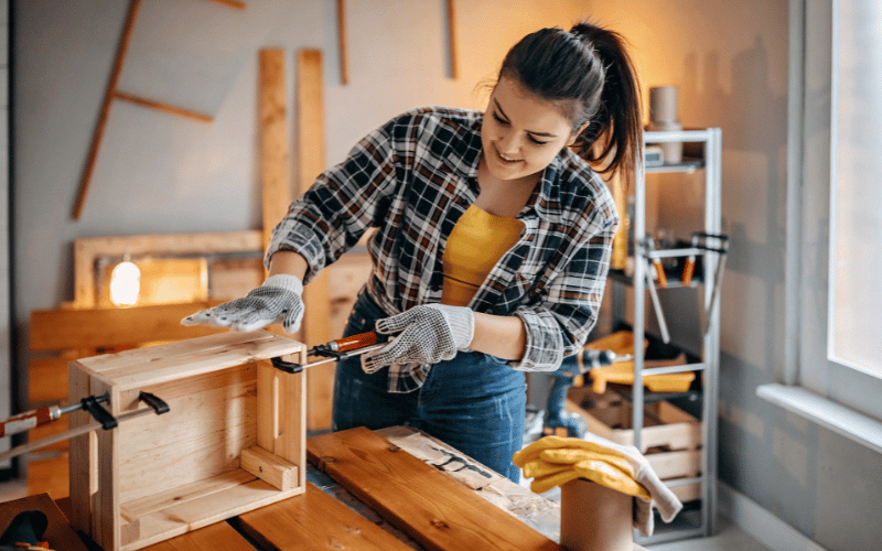 Young happy woman wearing jeans and plaid shirt and work gloves using tools to build wooden basket or storage container in home workshop