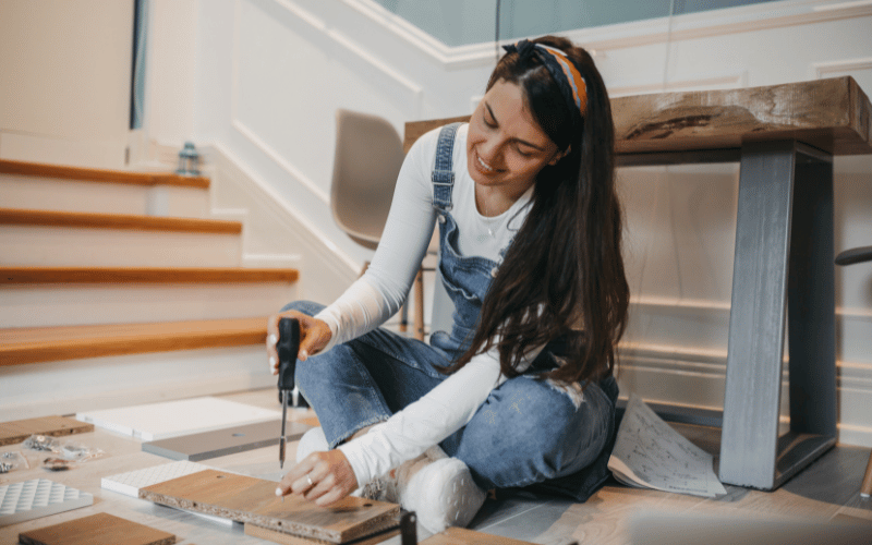 DIY young woman wearing overalls gently screwing in nails into small pieces of wood while sitting on floor next to cutting table 