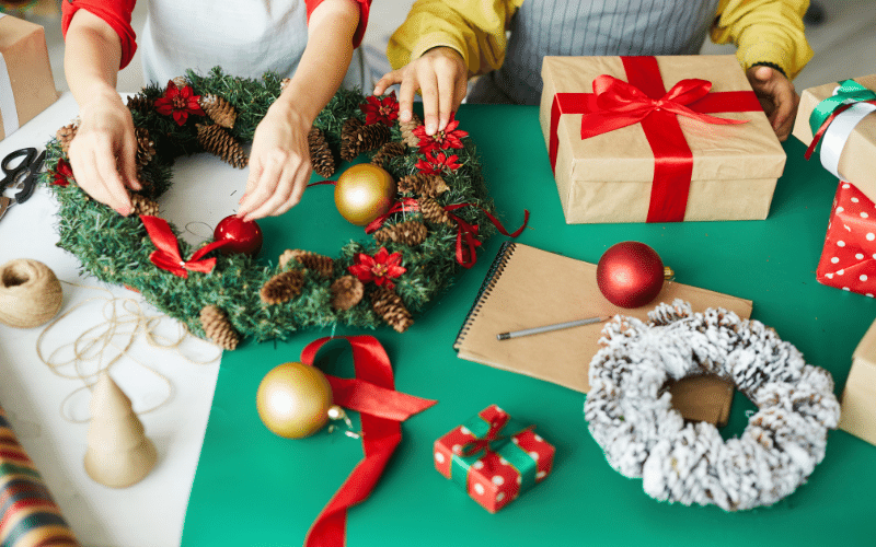 Making a Christmas Wreath over green construction paper on table. Red ribbon and gold bulbs