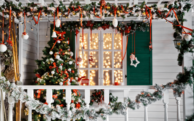 Front porch of white farmhouse decorated for Christmas with railing garland, dangling white stars on red ribbon and a decorated Christmas Tree