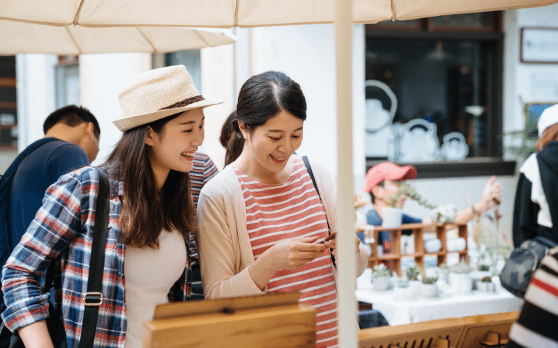 two ladies looking at a small object and smiling while at a market 