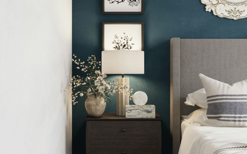 Dark green wall with home decor pieces like clock, lame and fake flowers on bedside table next to bed