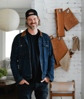 Celebrity Steve Ford smiling wearing black cap and jean jacket with his hands in his pockets in modern kitchen with exposed white brick in background