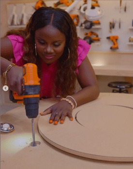 Celebrity Ati Williams wearing hot pink dress using orange electric drill on DIY wood project