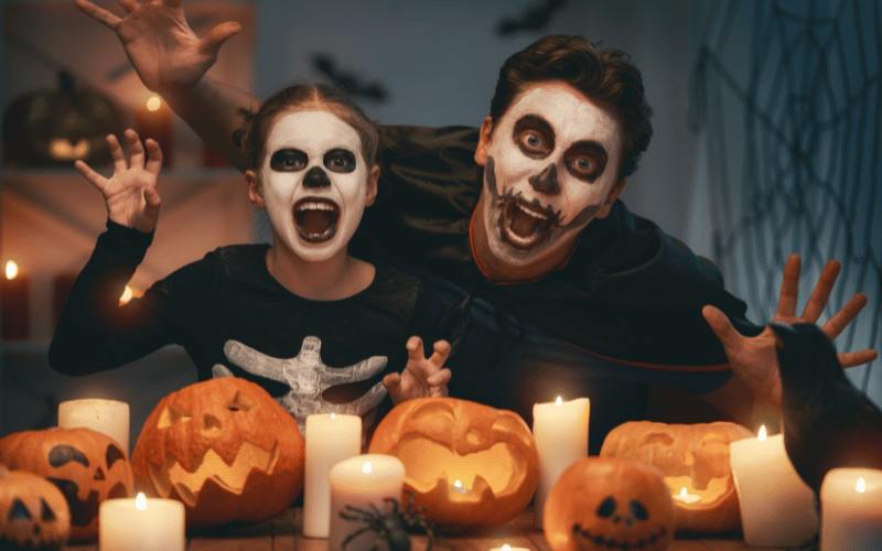 Daughter and Father dressed as skeletons wearing black and white face paint in front of carved pumpkins and white candles making a face