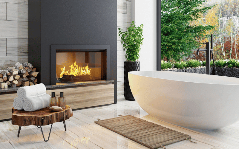 Large deep soaker tub with open window and forest, large tall plant, fireplace on black wall and spa towels and lotions on a wooden trunk table