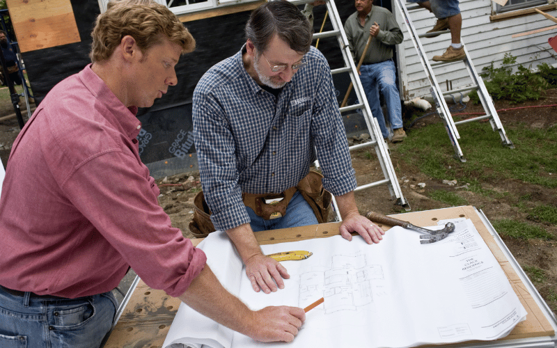 PBS' Kevin O'Connor and another worker looking at blueprints for a house rebuild on a table outside