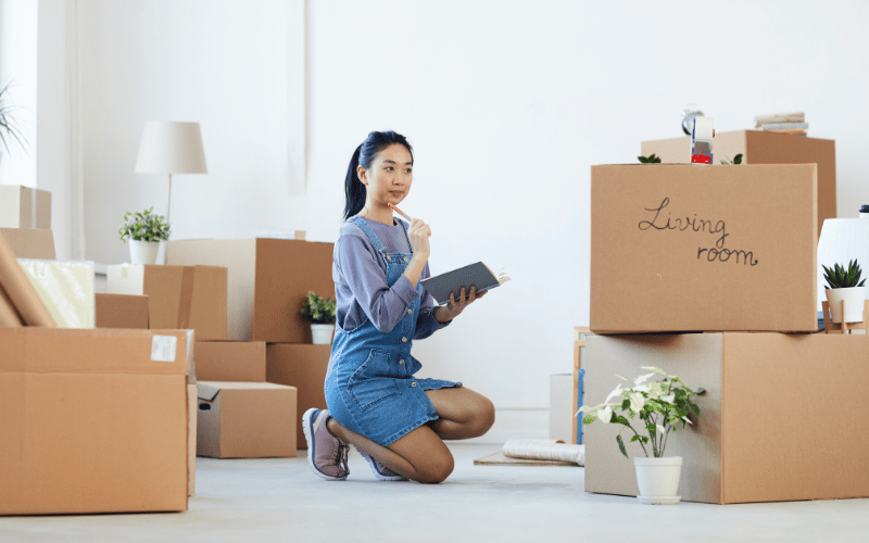 Young Asian woman wearing jean overalls kneeling on floor looking at boxes