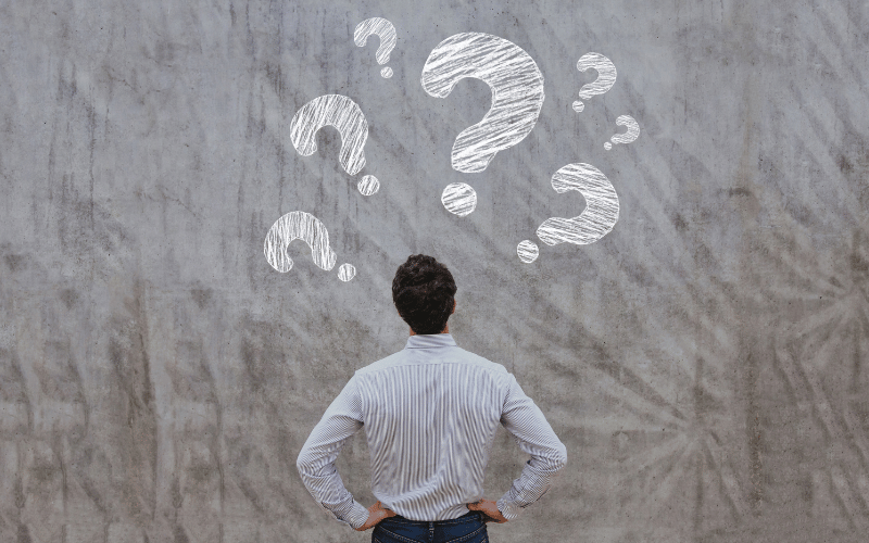 Man with black hair from back wearing striped dress shirt and hands on hips, looking at concrete wall with white chalk question marks