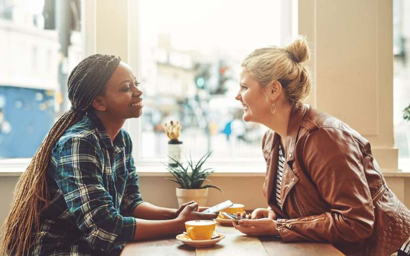 Two young women friends smiling and laughing together over coffee sitting in coffee shop at wooden table in window