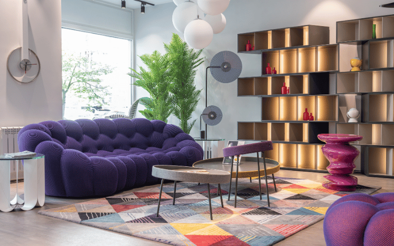 Purple maximalist room with purple couch and chair and lighter purple sculpture and abstract circular tables next to full wall open concept shelving