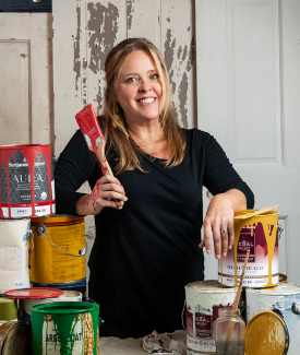 Ashley Morrill Ashley Morrill standing in a room holding a paintbrush surrounded by large paint cans smiling