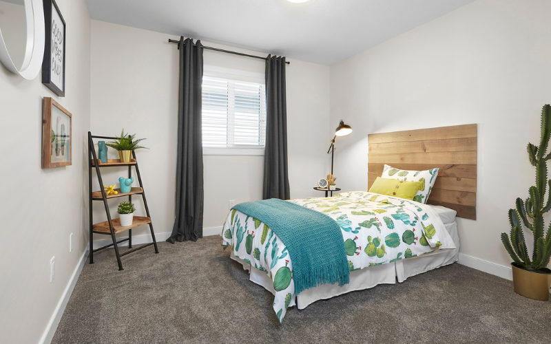 Small bright bedroom with wooden wall mounted headboard and white bedding with cactus design on it. Opposite bed on left is three level black metal and wood rack containing plants beside black curtains open window bright day outside.